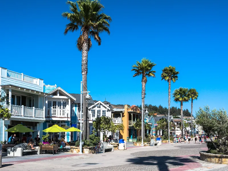 Palm trees line a sunny street with colorful buildings and people walking and sitting under umbrellas.