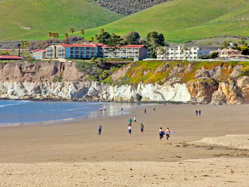 People walking and carrying surfboards on a sandy beach with cliffs and buildings in the background.