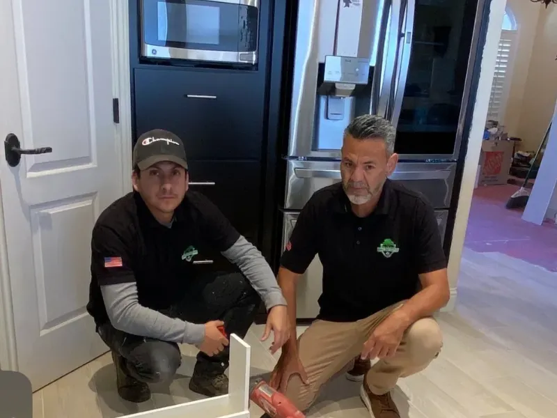 Two men in work uniforms crouch on a kitchen floor near a refrigerator and a power tool.