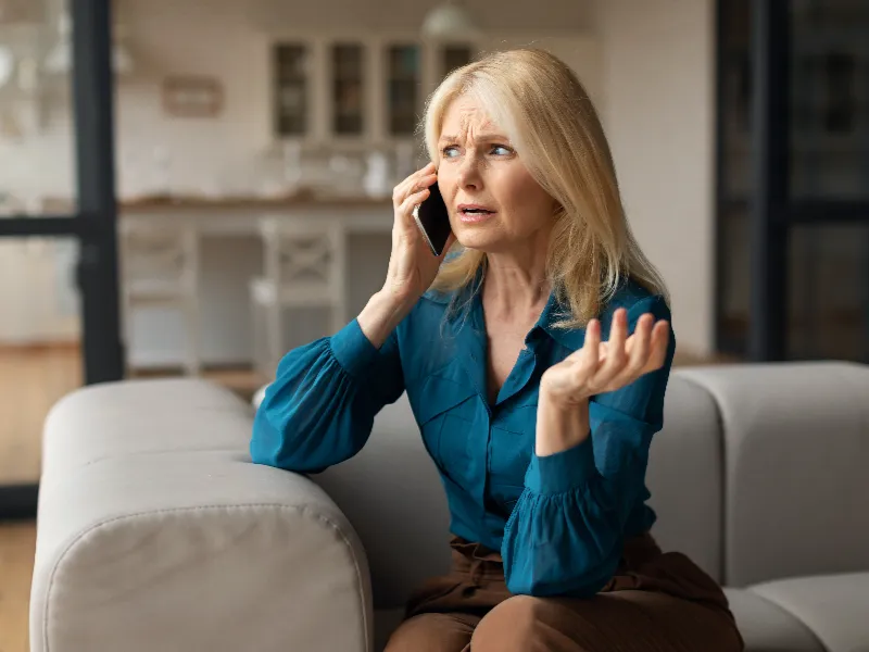 A woman with blonde hair looks worried while talking on a phone sitting on a couch.