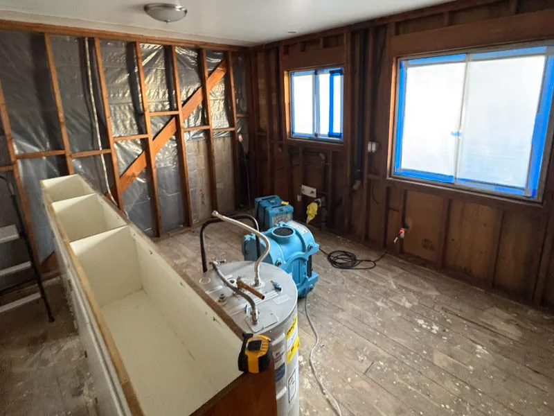 Room under renovation with exposed wooden studs, a water heater, and blue drying equipment on a wooden floor.
