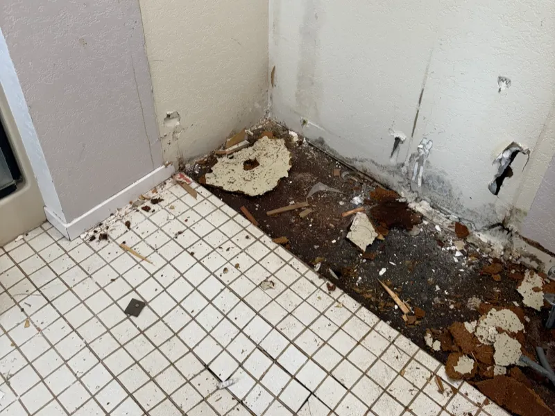 Section of tiled floor with damaged wall and debris scattered along the baseboard area.