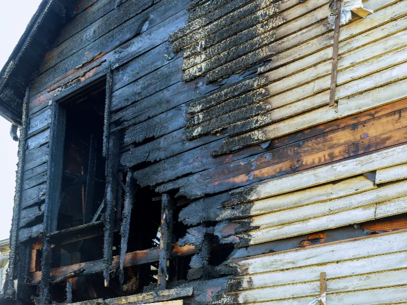 Partially burned wooden exterior wall of a house with a charred window frame.