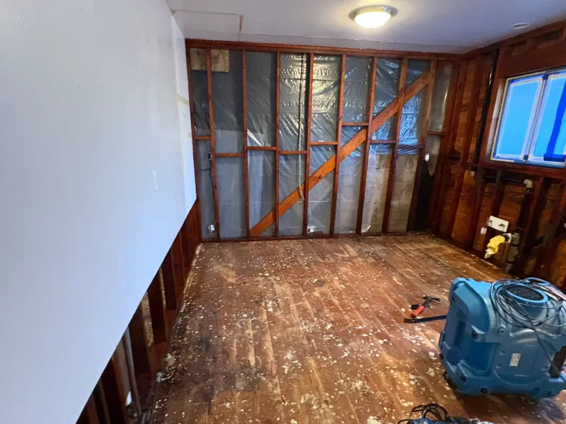 Room under renovation with exposed wall studs, a bare wooden floor, and a blue air mover on the floor.