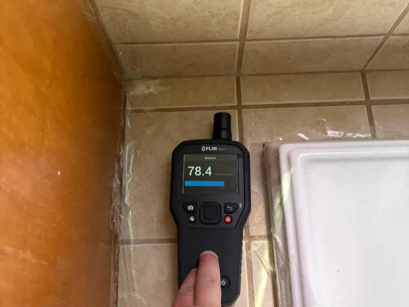 Hand holding a FLIR MR77 moisture meter displaying a reading of 78.4 on tiled floor near a wooden surface.