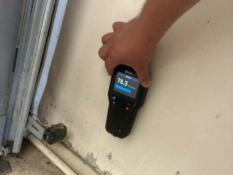 Hand holding a moisture meter showing a reading of 76.3 on a beige wall.
