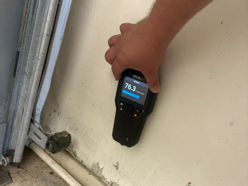 Hand holding a moisture meter showing a reading of 76.3 on a beige wall.