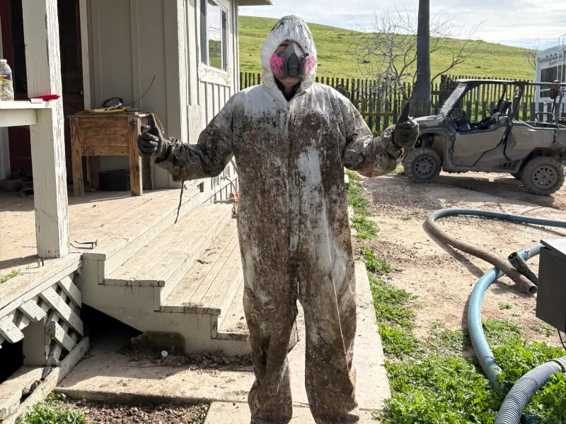 Person in a dirty protective suit and respirator giving thumbs up outside a building.