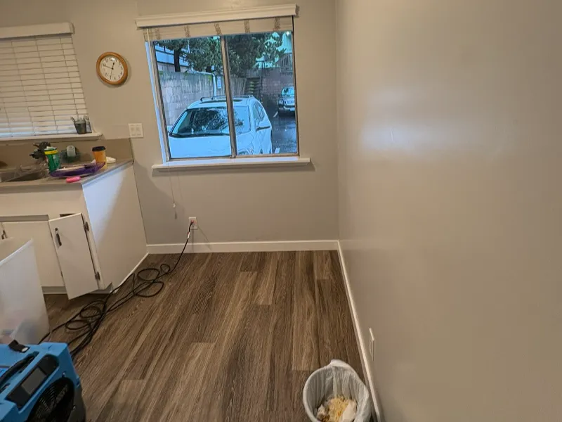 Empty kitchen corner with wood flooring, a window showing parked cars, and a trash can with food waste.