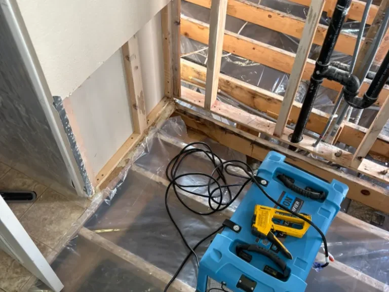 Blue air mover and yellow moisture meter on plastic-covered floor in a partially demolished room.