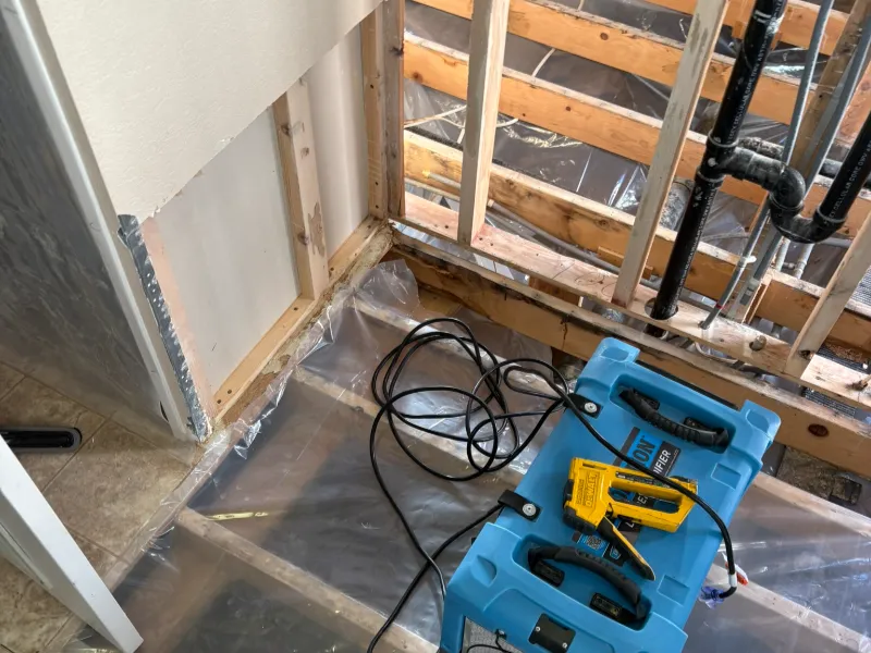 Blue air mover and yellow moisture meter on plastic-covered floor in a partially demolished room.