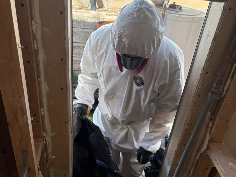 Person in white hazmat suit and respirator holding a black bag in a doorway.