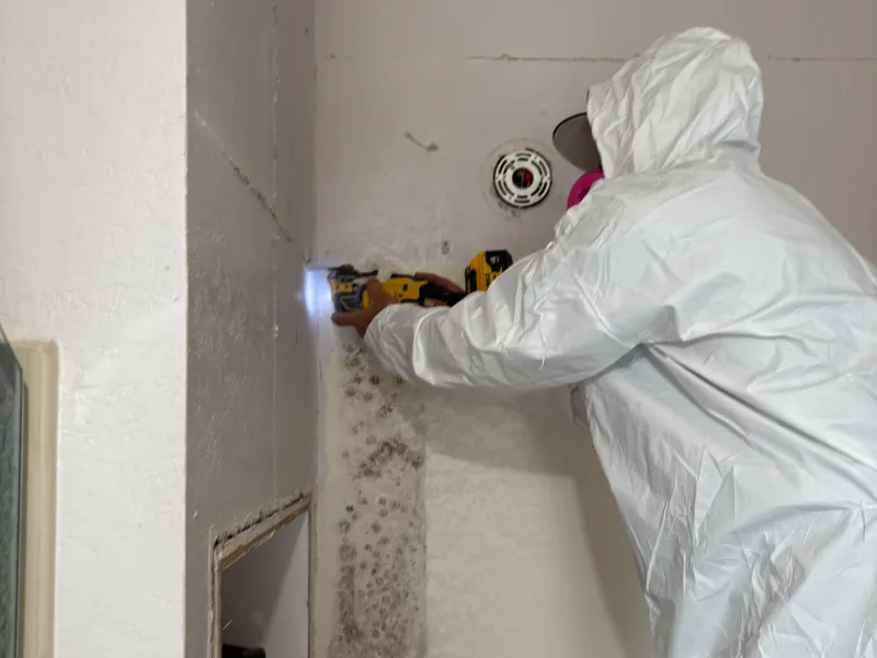 Person in protective suit using a drill on a wall with visible mold.